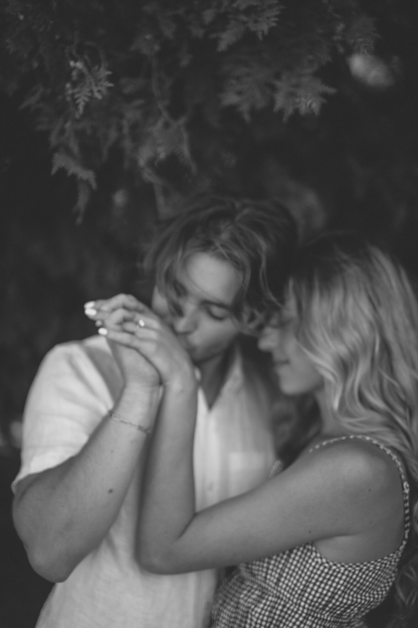Two women face each other closely, holding hands, with a dark background and foliage above them.