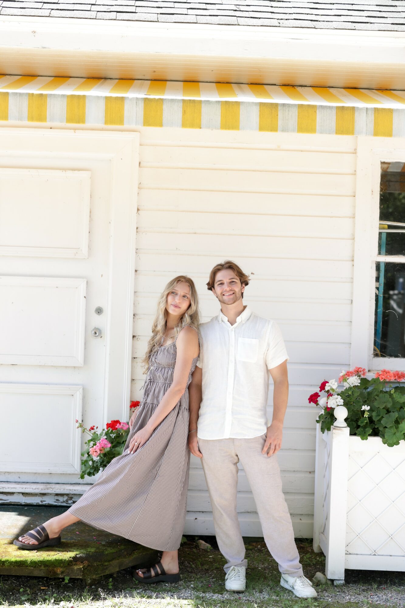 Two people standing outside a white building with yellow striped awning and flower planter, smiling at camera.