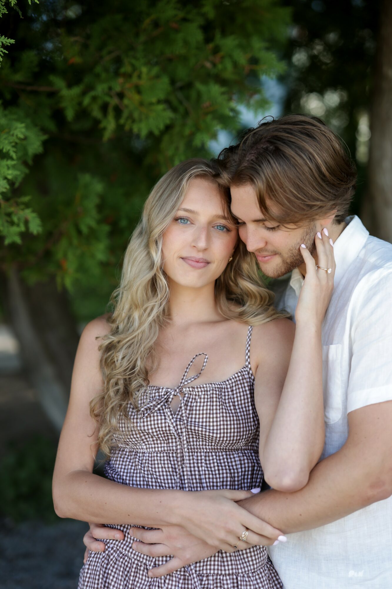 A young couple standing close outdoors, with trees in the background, embracing and looking content.