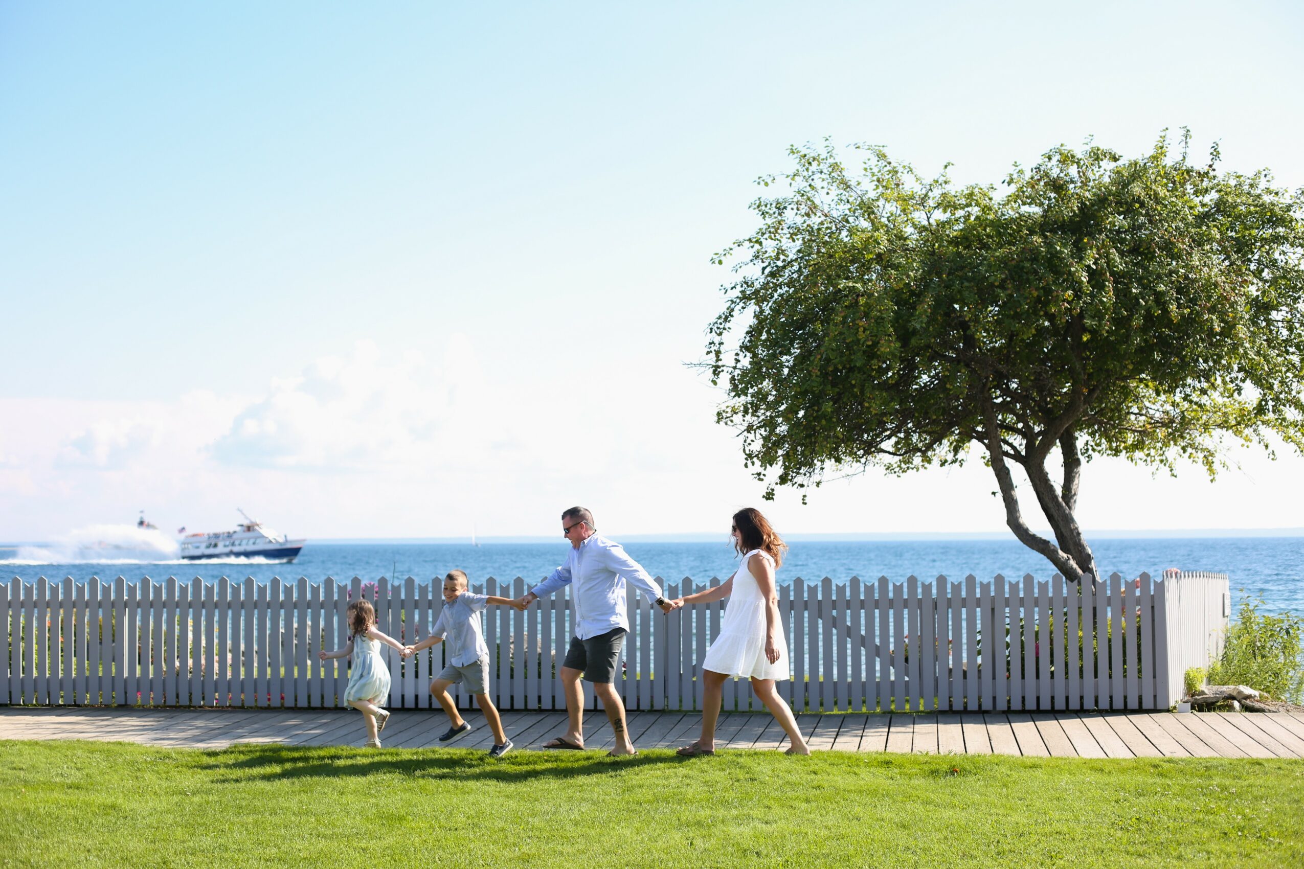 Family of four walking along a wooden path near a white fence and a tree, with water and a ship in the background.