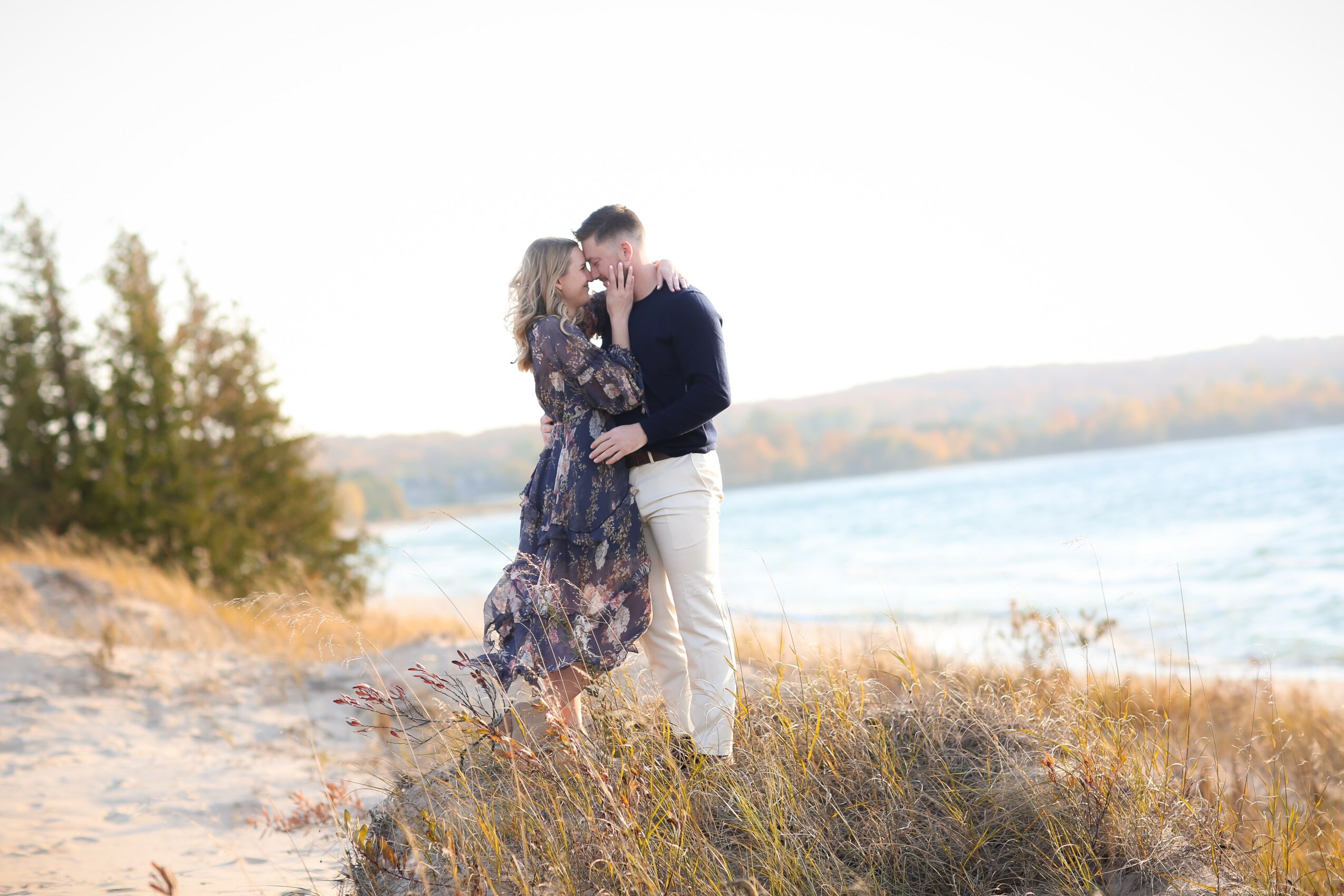 A couple stands on a grassy shoreline near water, embracing and touching foreheads, with trees and water in the background.