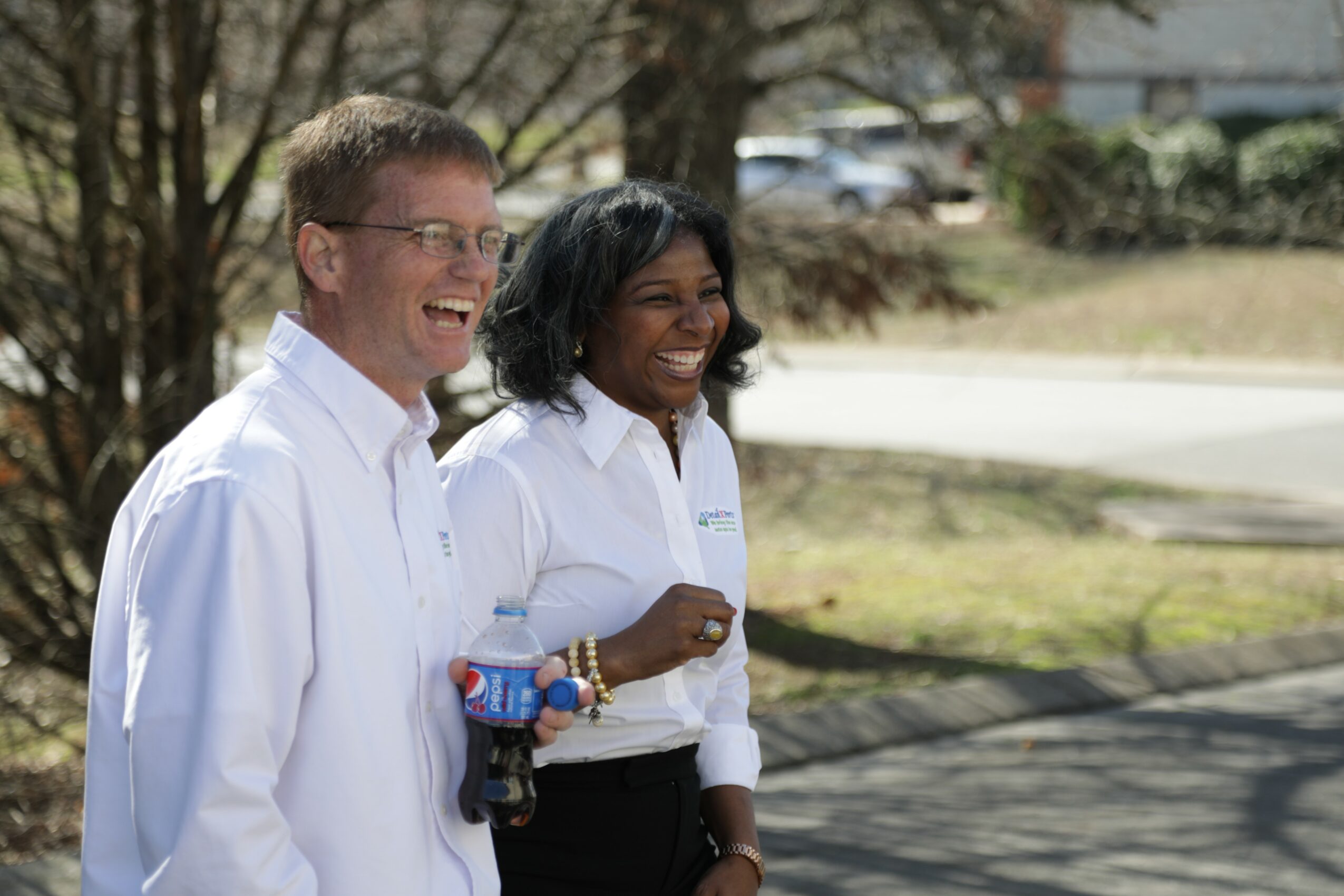 Two smiling women in white shirts standing outdoors with trees and a road in background.