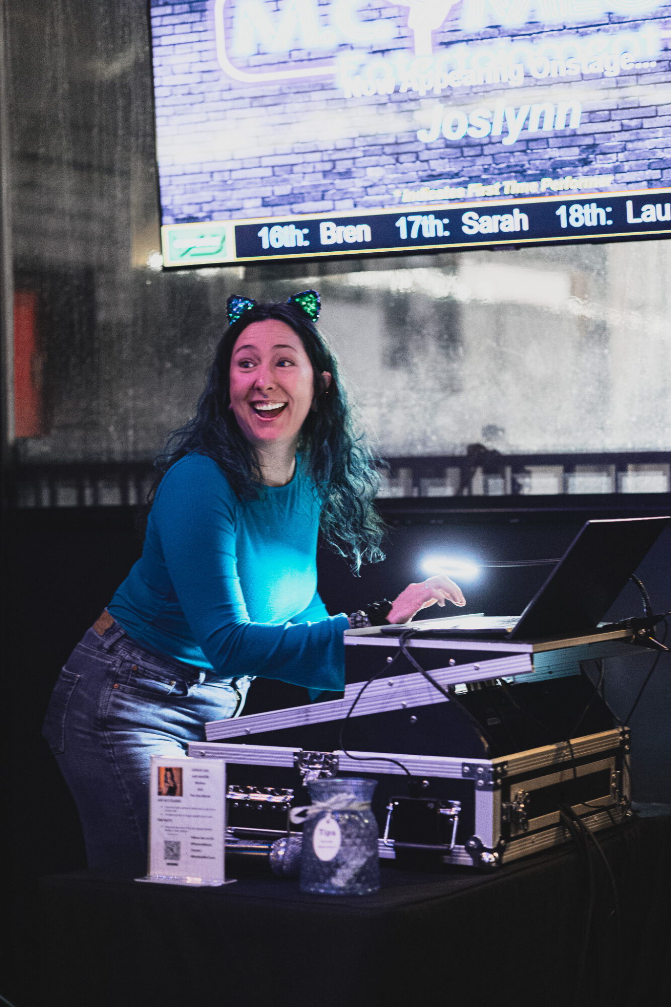 Woman with long dark hair smiling, wearing a blue top, standing behind DJ equipment in a dimly lit setting.