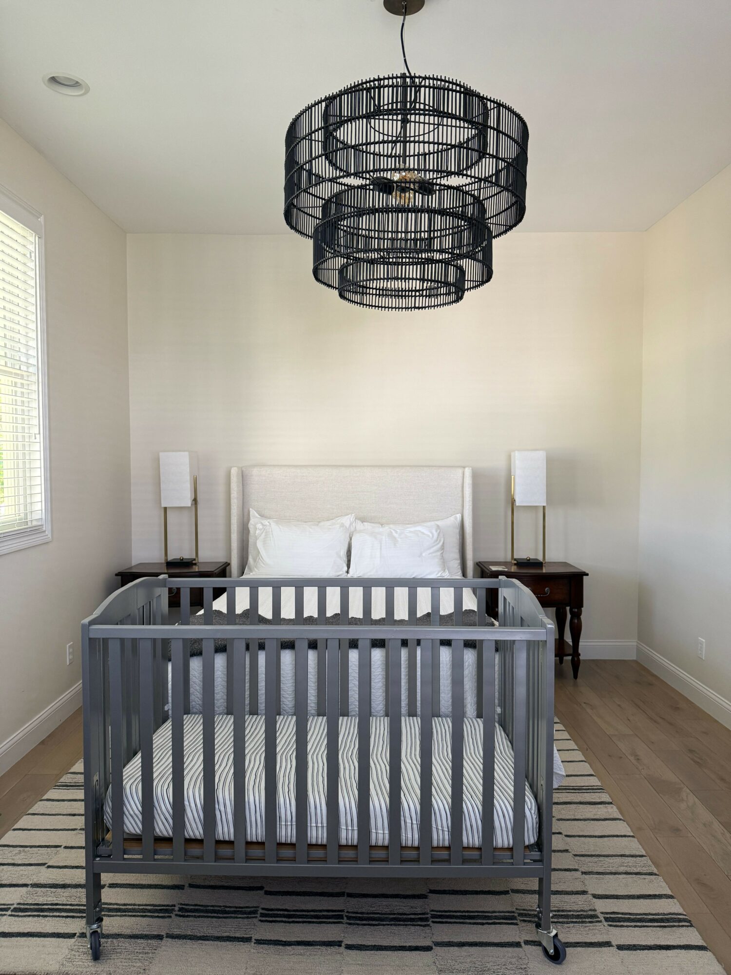 Bedroom with crib, bed, two nightstands, lamps, and a black chandelier, with a window on the left wall.