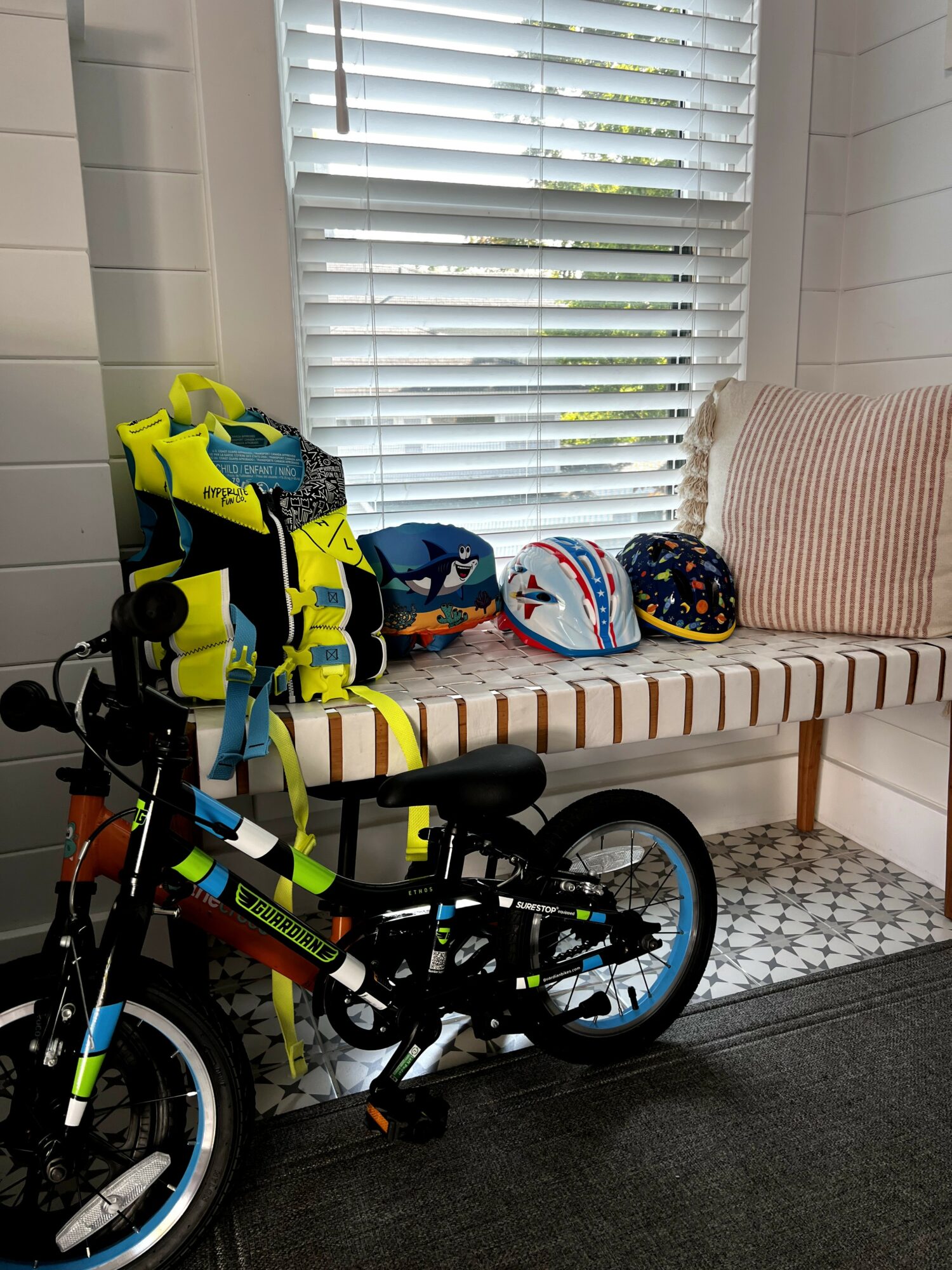 Indoor scene with a bicycle, helmets, and a backpack on a striped bench near a window with blinds.