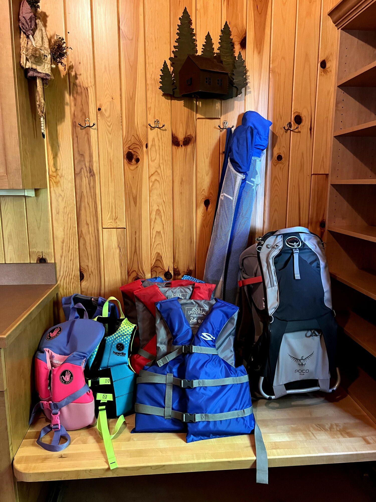 Stacked backpacks and bags on a wooden table in a room with wooden panel walls and a shelf.