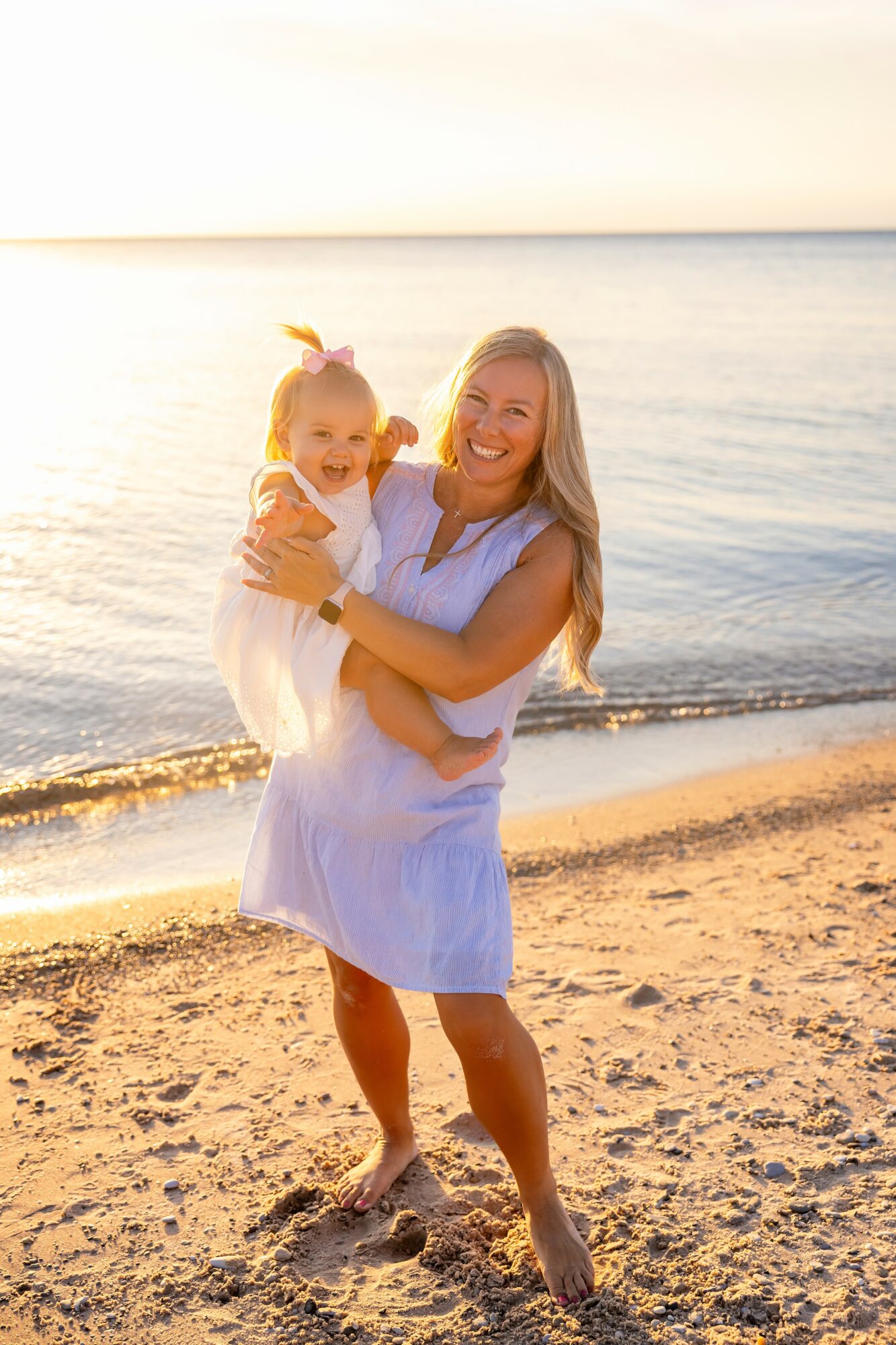 Woman holding a child on a beach at sunset, smiling, with water and sand in background.