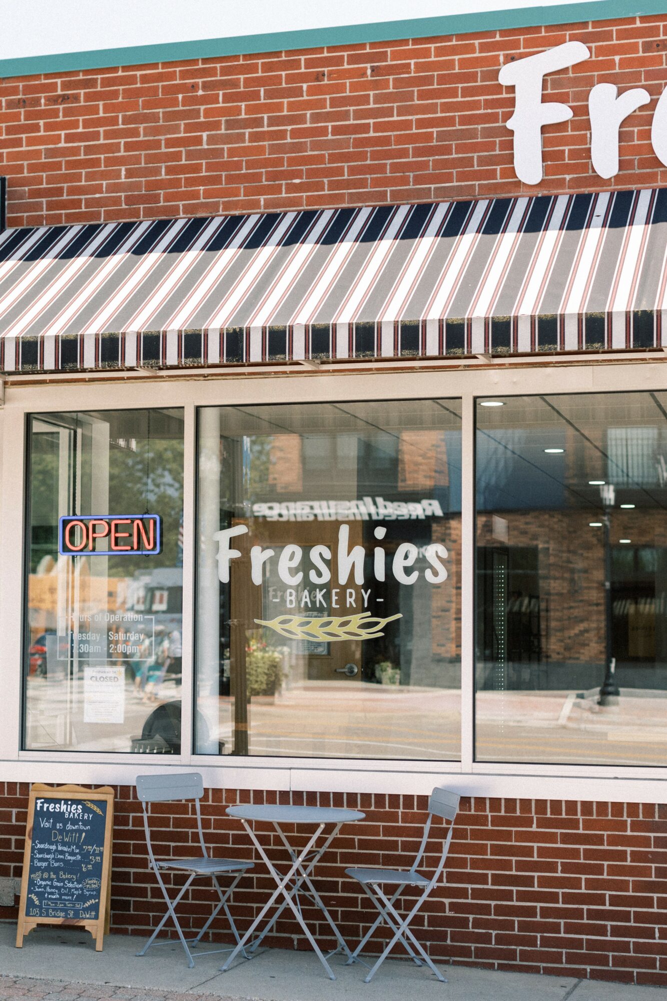 Bakery storefront with striped awning, glass windows, and outdoor seating with table and chairs.