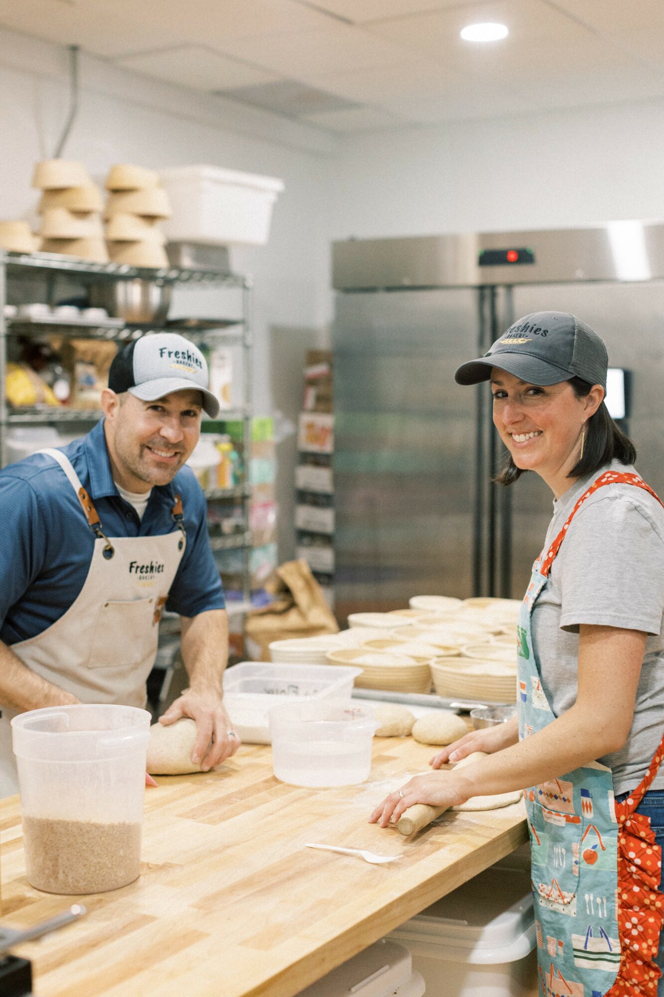Two people smiling and working at a bakery counter with dough and baking supplies.