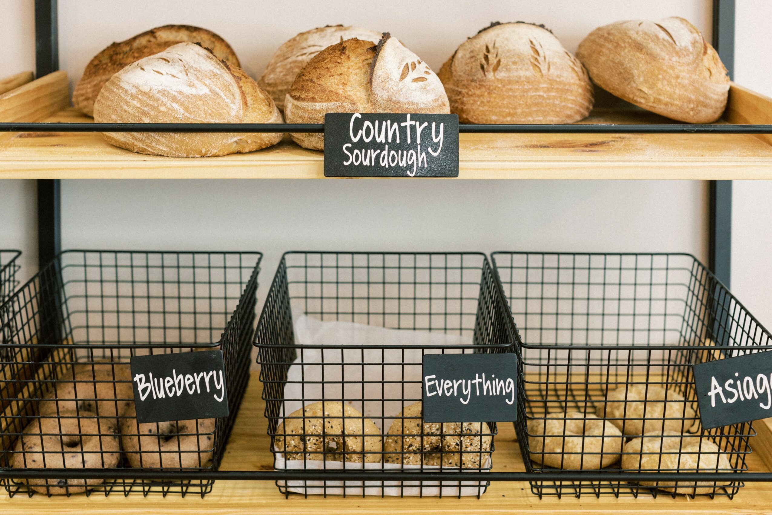 Three wire baskets filled with potatoes, labeled 'Blueberry,' 'Everything,' and 'Asian,' on a shelf with bread loaves above.