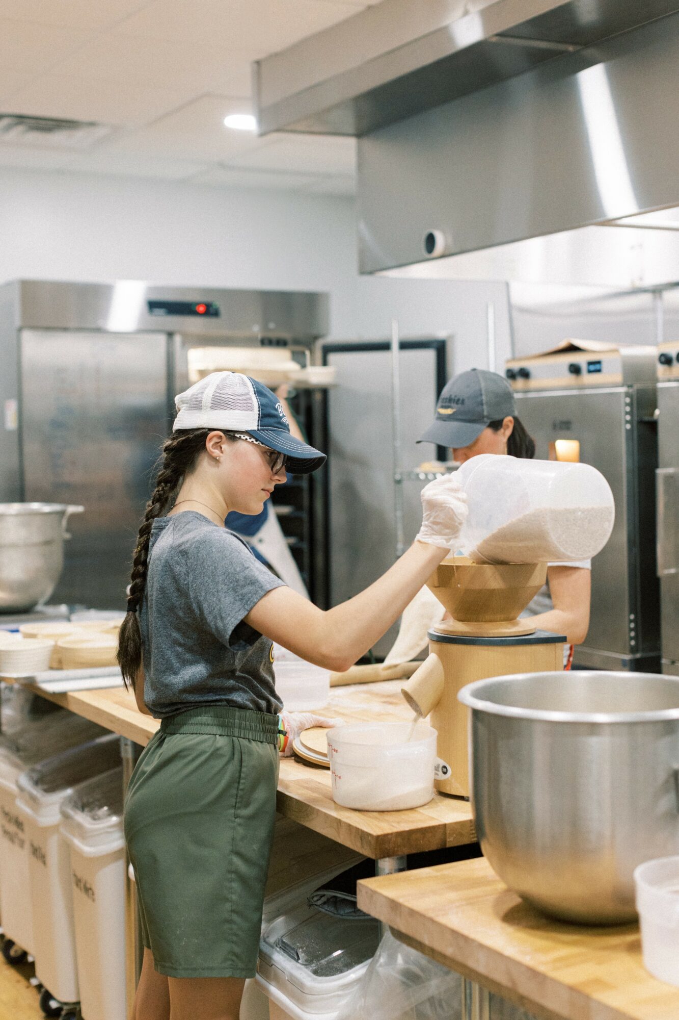 Two people working in a commercial kitchen with stainless steel appliances and wooden countertops, handling baking supplies.