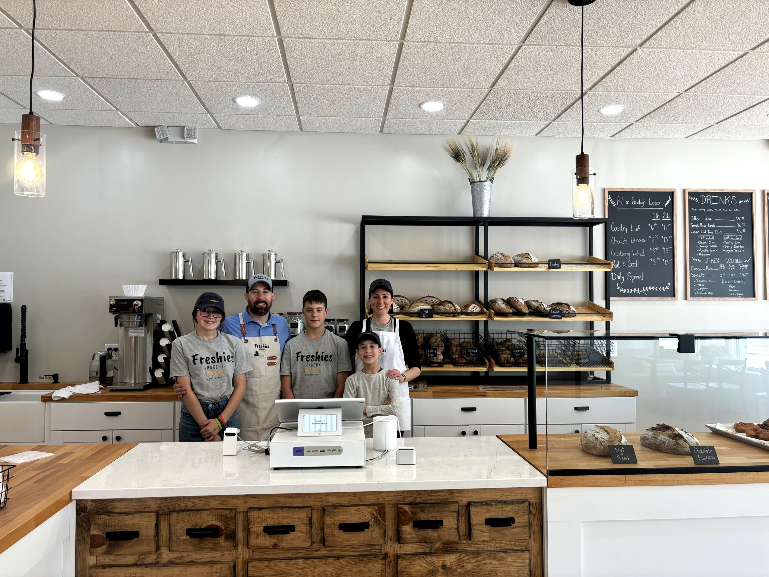 Four staff members stand behind a counter in a bakery or cafe, with shelves of bread and chalkboard menus behind them.