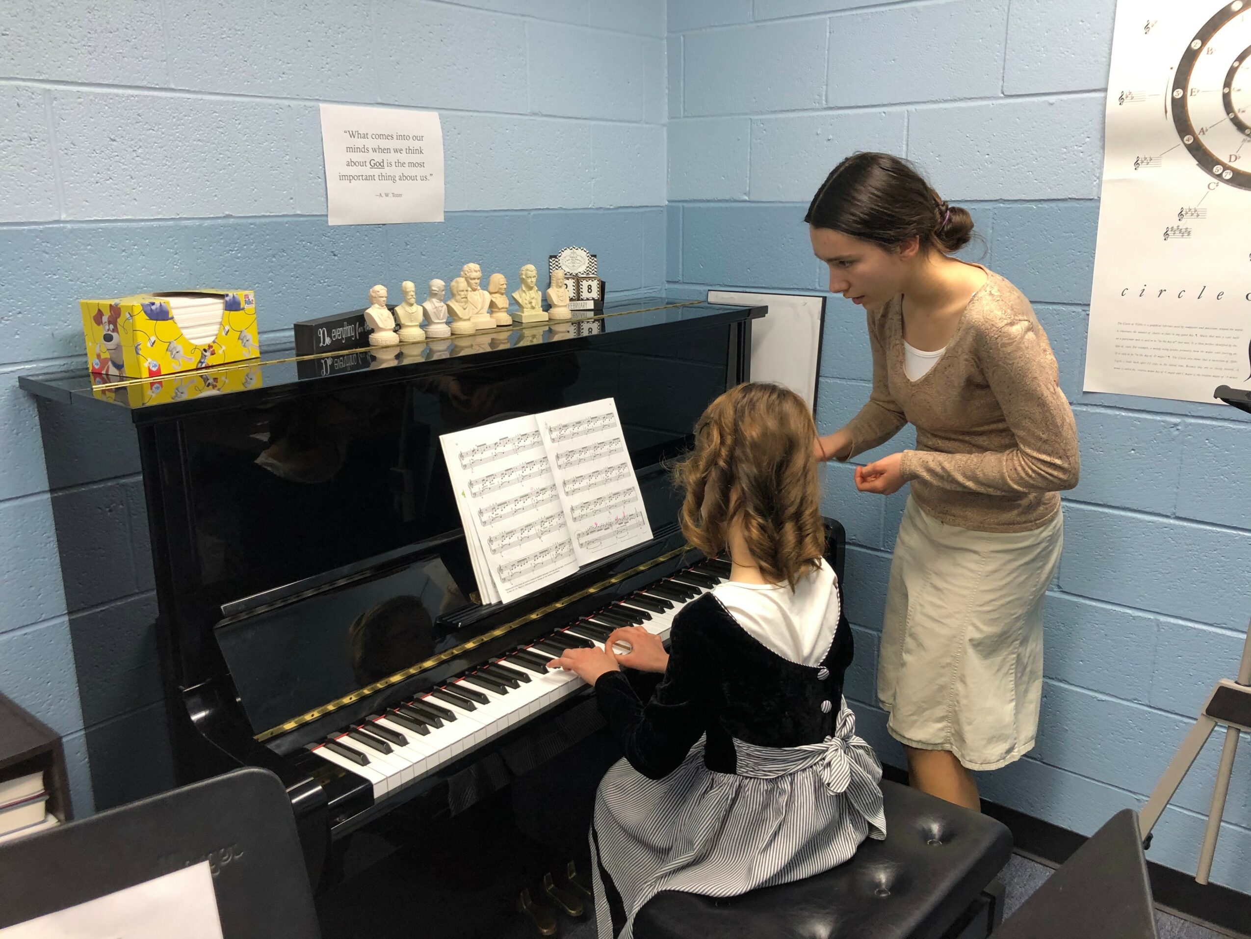 A woman and a girl are at a piano in a room with blue walls, the girl is playing, the woman is instructing.