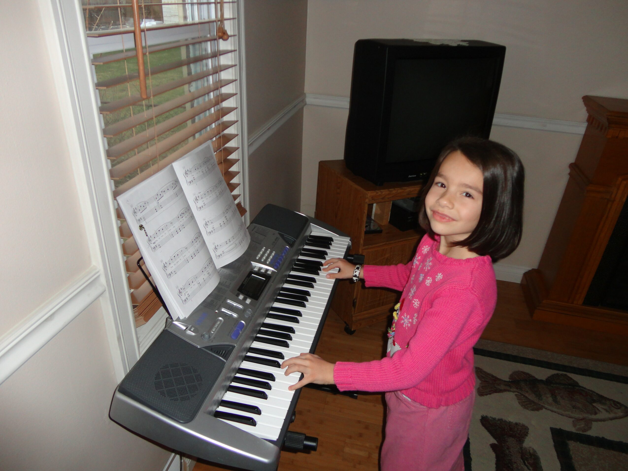 Young girl in pink sweater playing a keyboard piano indoors, smiling, with a TV and window in background.