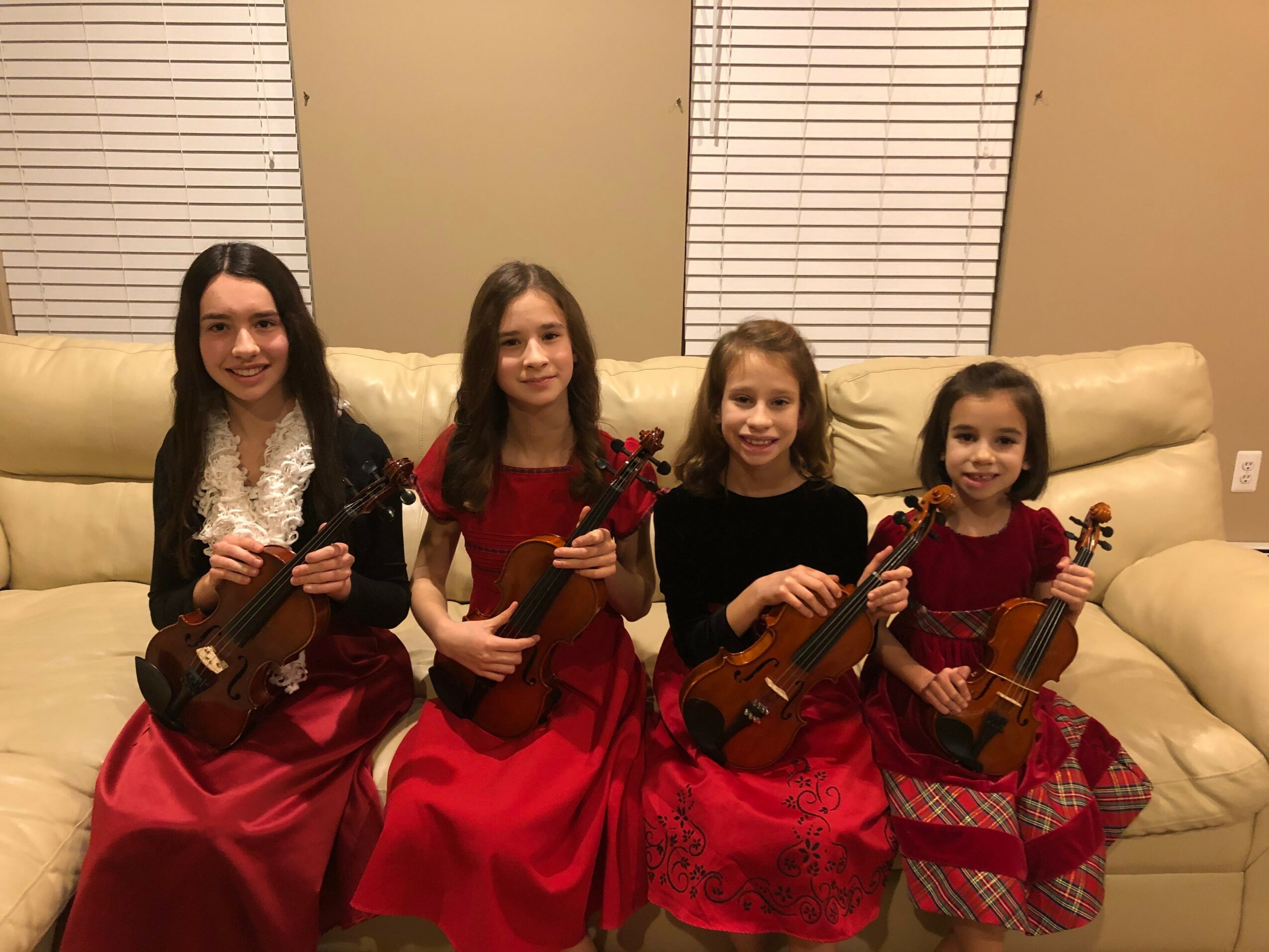 Four young girls in red dresses sitting on a beige couch, each holding a violin, smiling at the camera.