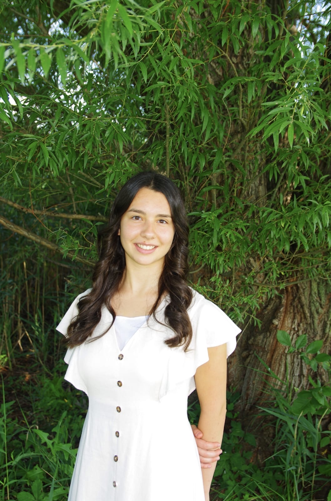 Young woman with long dark hair smiling outdoors in front of green foliage, wearing a white dress.