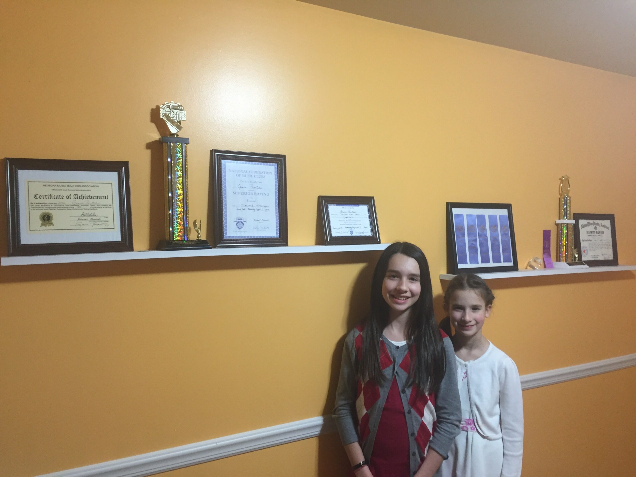 Two girls stand in front of a yellow wall with framed certificates and trophies on a shelf.