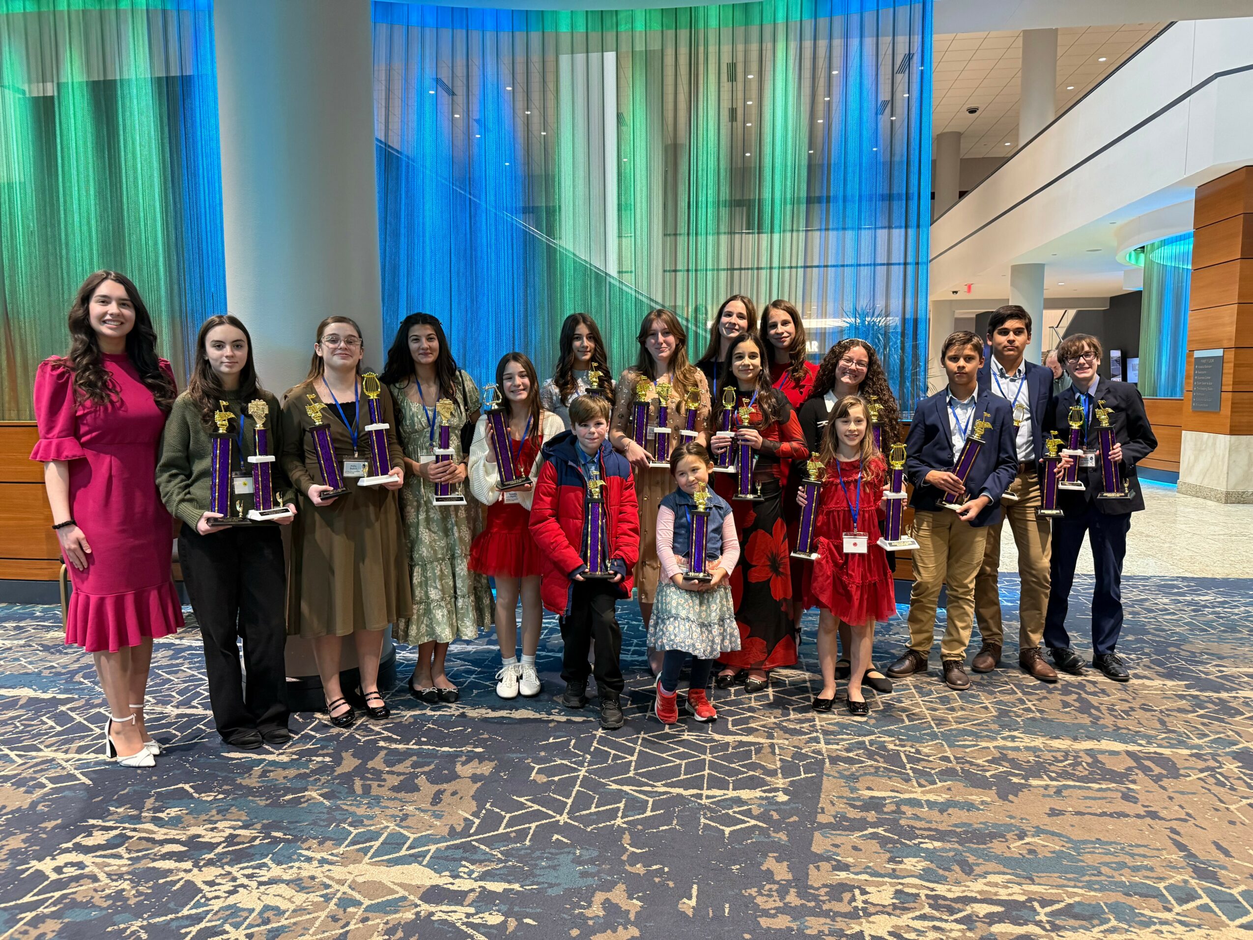 Group of children and adults standing in a hotel lobby holding awards, colorful curtains in the background.