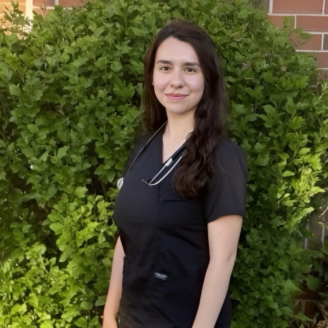 Young woman with long dark hair wearing a black uniform and stethoscope, standing outdoors in front of green bushes.