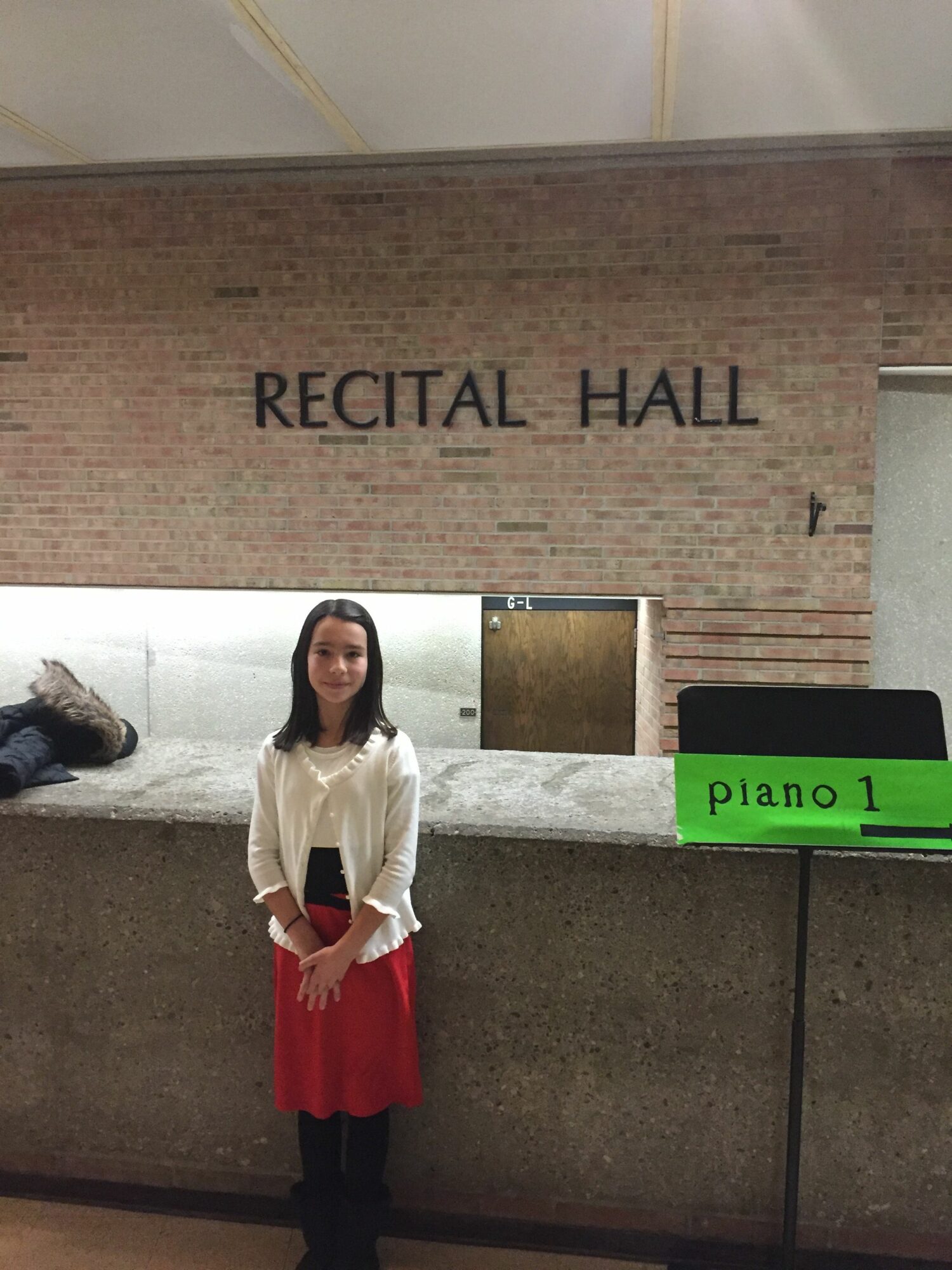 Girl standing in front of a brick wall with 'RECITAL HALL' sign, wearing a white top and red skirt.