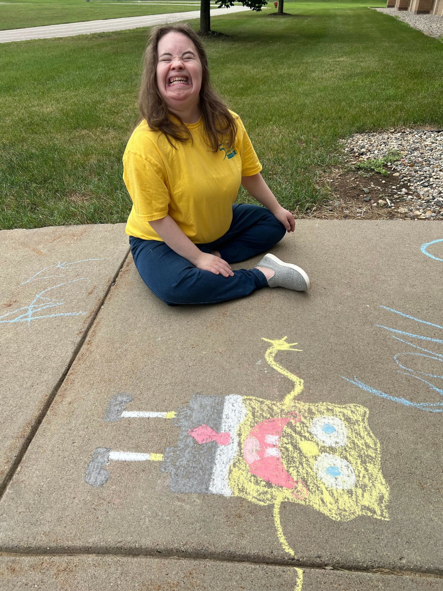 Child sitting on sidewalk smiling, wearing yellow shirt and blue pants, with colorful chalk drawing of a character nearby.