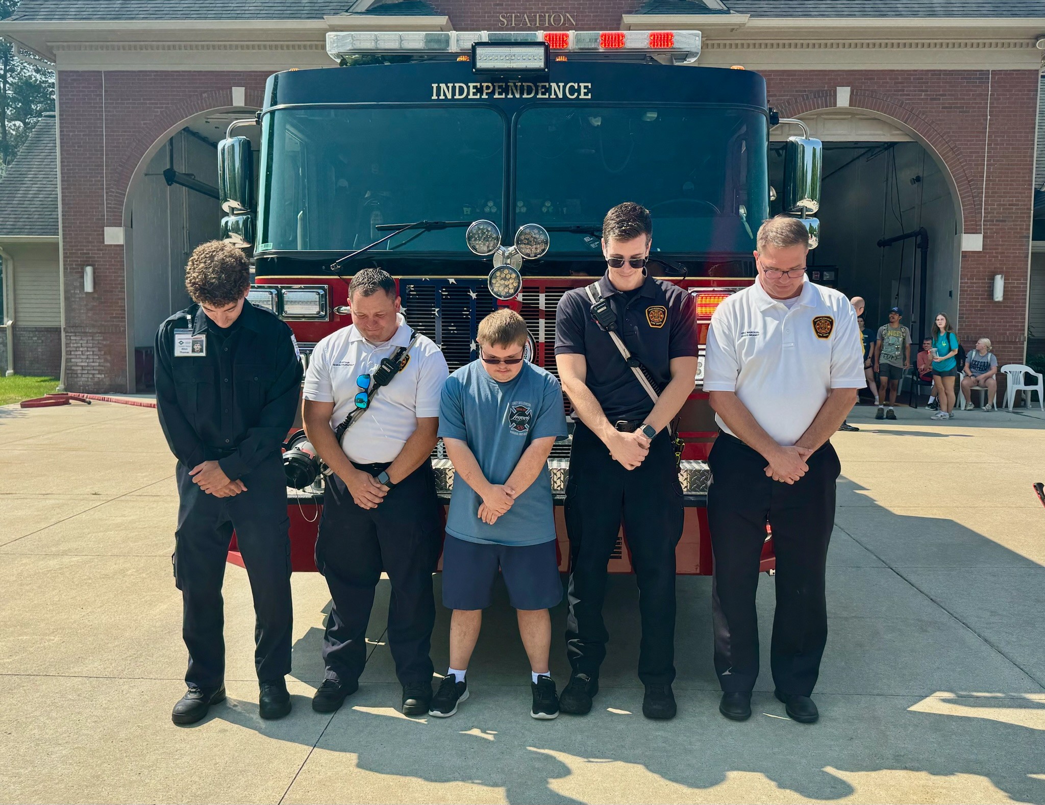 Five people standing in front of a large fire truck, with a brick building in the background.