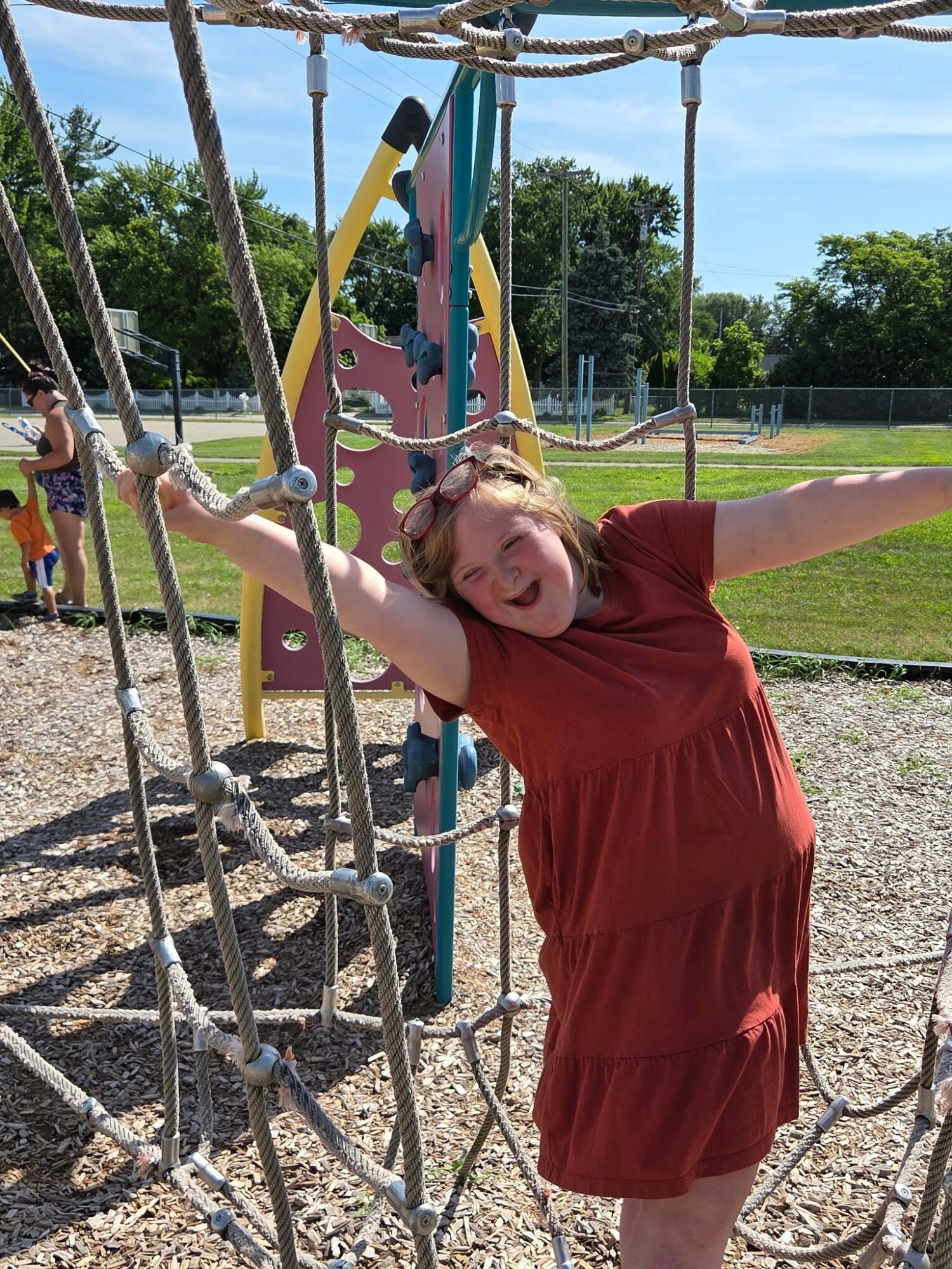Girl in red dress smiling with arms outstretched on playground climbing structure outdoors.