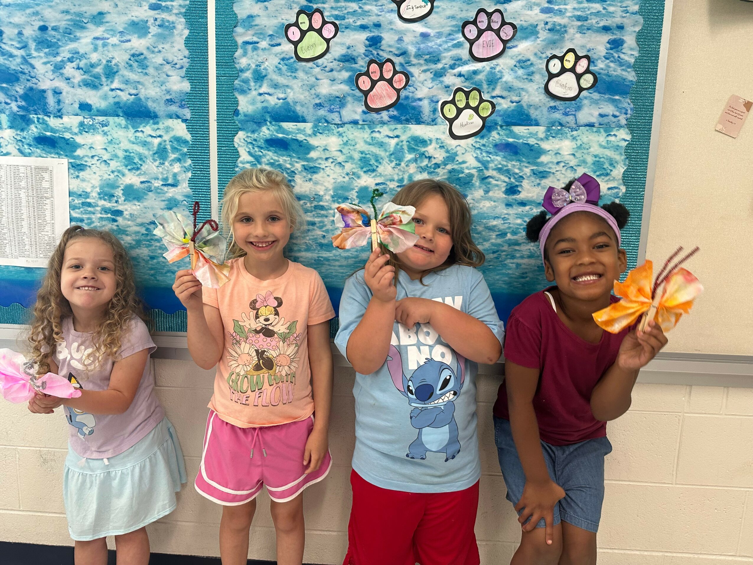 Four children stand in front of a blue wall decorated with paw prints, holding crafts and smiling.