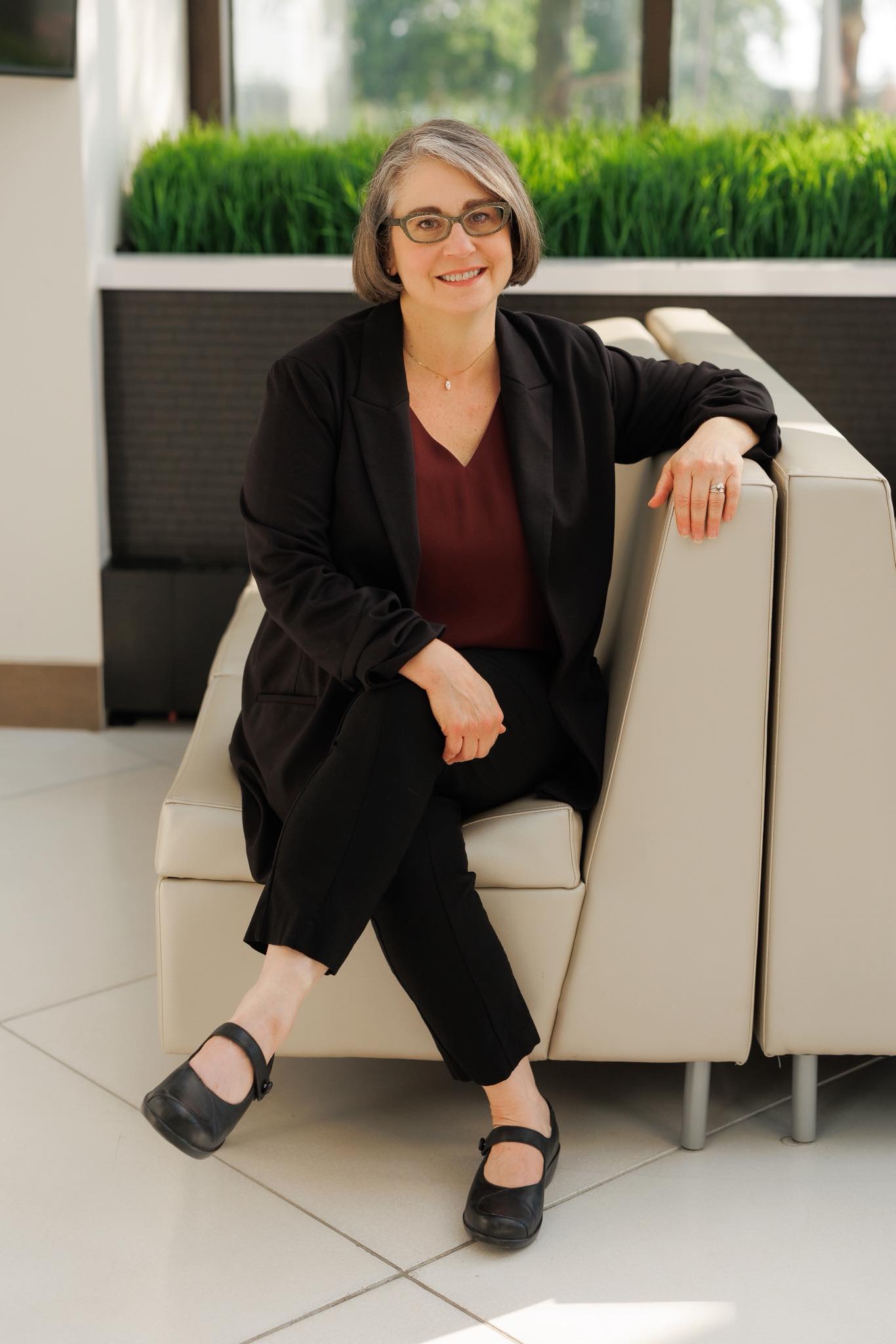Woman with glasses and short hair sitting on a white chair, smiling, in a modern indoor setting with greenery outside.