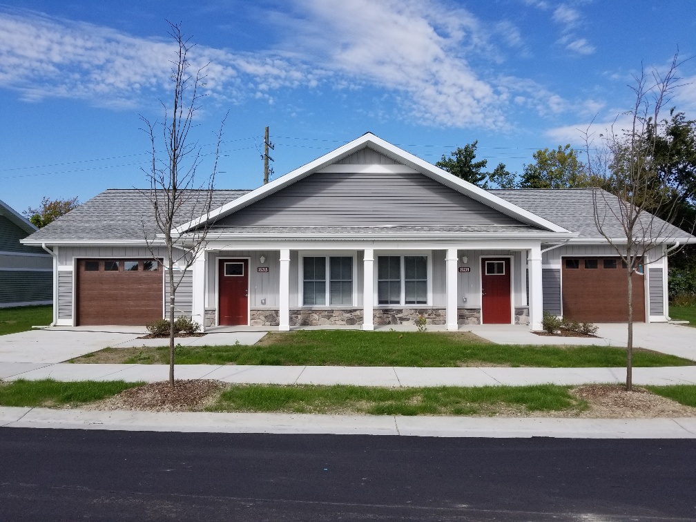 Single-story building with three front doors, two garage doors, and a central porch with three windows, trees, and a sidewalk.