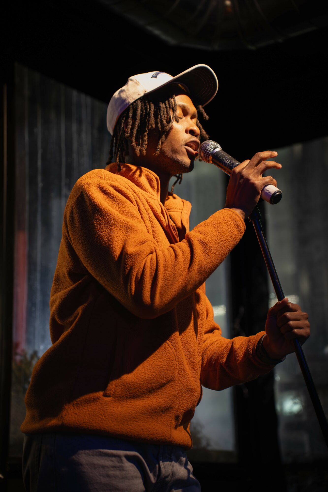 Person singing into a microphone on stage, wearing a white cap and orange hoodie.