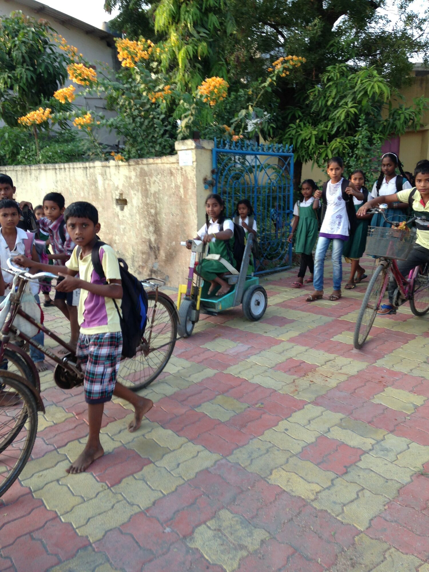 Group of children with backpacks and bicycles on a paved street, some walking and some riding bikes, in a neighborhood.