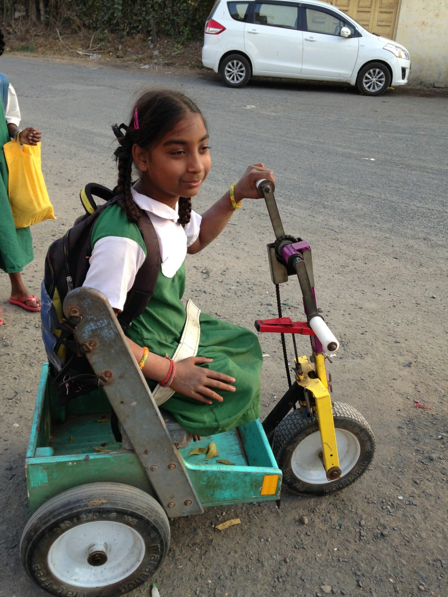 Young girl sitting on a green and yellow tricycle with a black backpack, smiling outdoors near a white car.