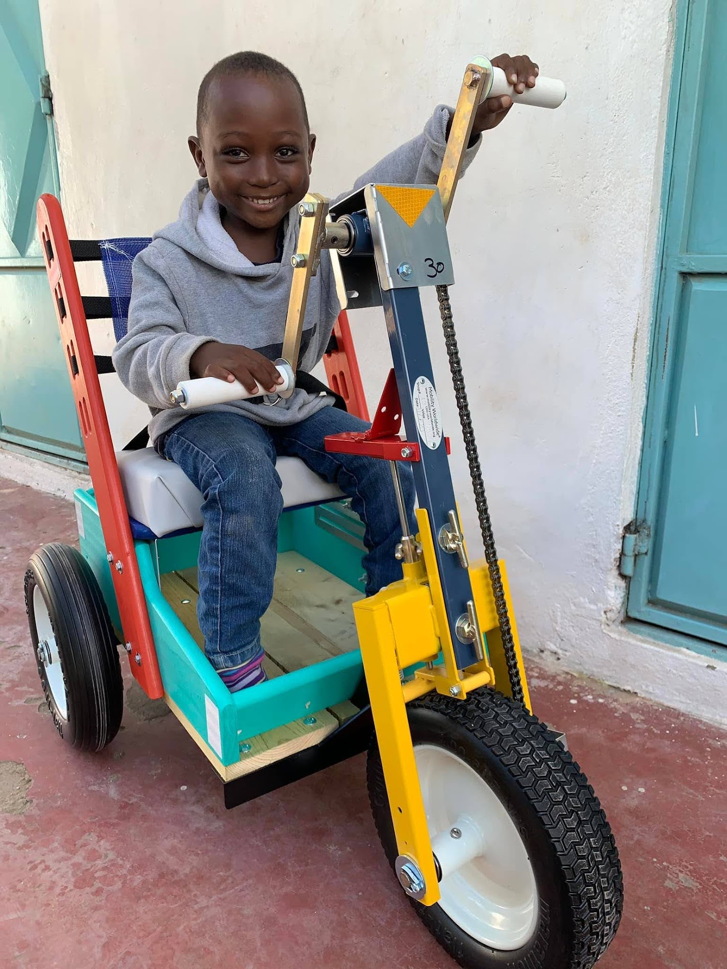 Child sitting on a colorful homemade bicycle with a large front wheel, smiling, holding a remote control, outdoors.
