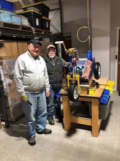 Two men standing next to a yellow and black machine in a warehouse, surrounded by shelves and storage boxes.