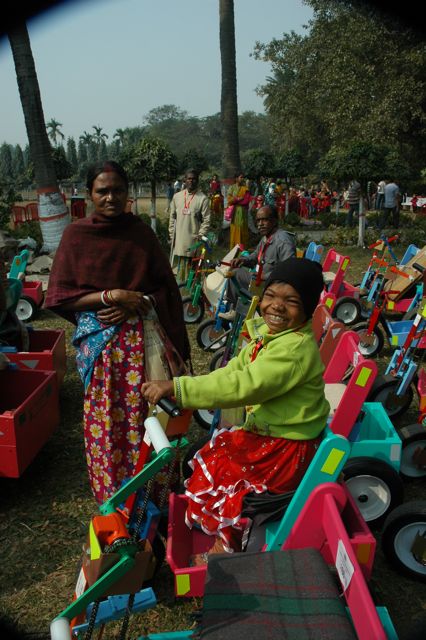 Child in bright green jacket and red skirt sitting in colorful toy car, smiling, outdoors with adults and trees in background.