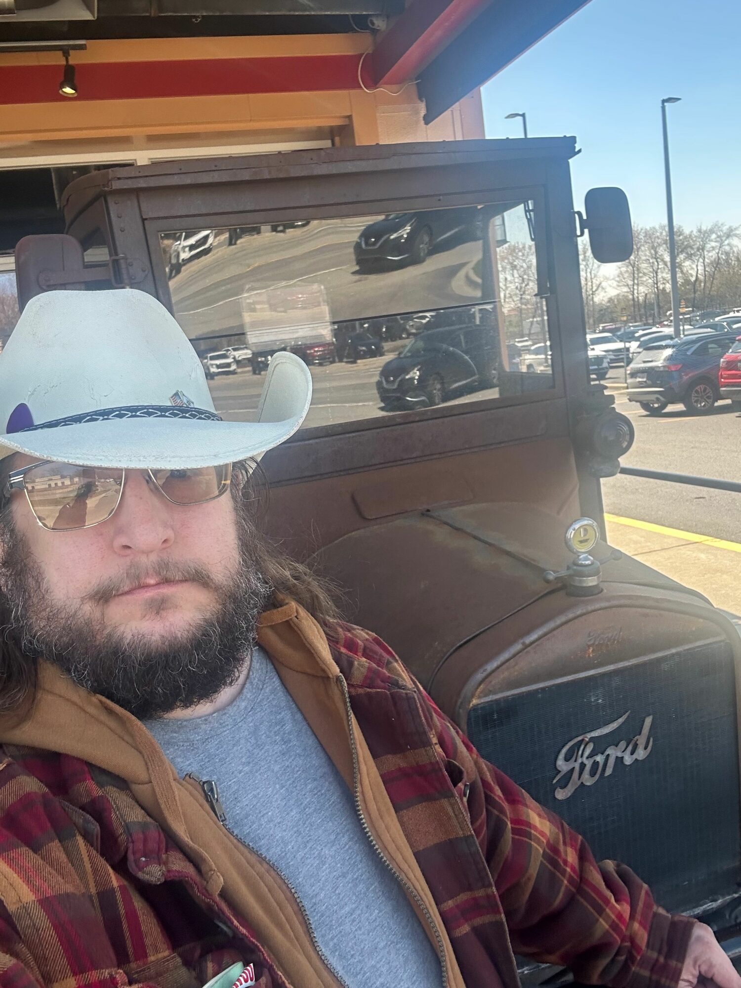 Man with beard wearing sunglasses and a cowboy hat, sitting in front of a vintage Ford vehicle outside a store.