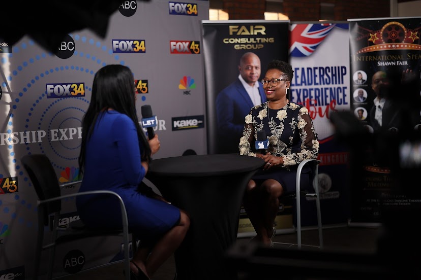 Two women seated at a table during an interview or discussion, with a backdrop of banners and logos behind them.