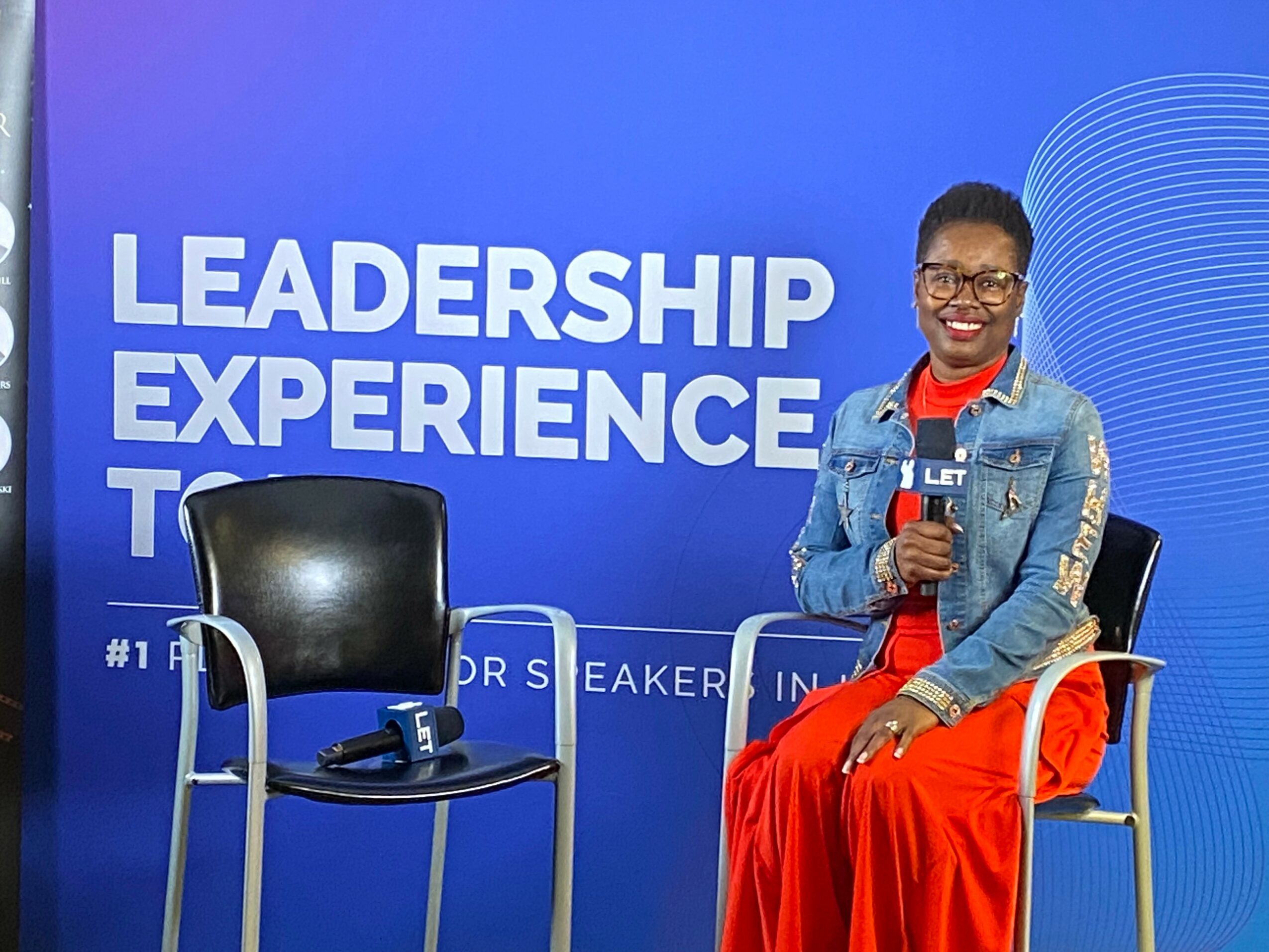 Woman sitting on a chair holding a microphone, smiling, in front of a blue backdrop with text.