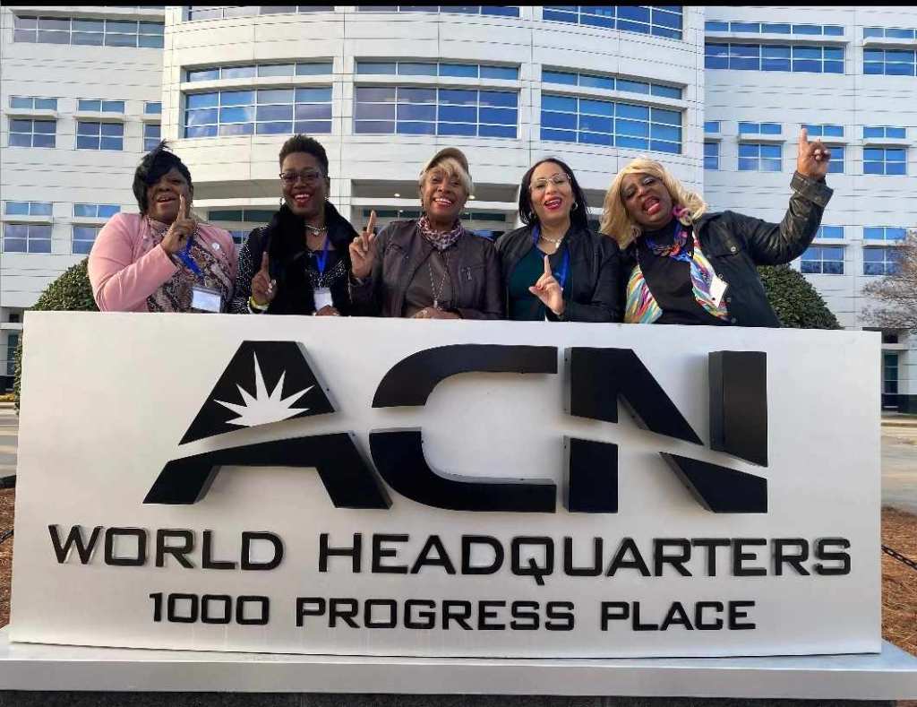 Five women standing behind a sign that reads 'ACN World Headquarters, 1000 Progress Place,' smiling and waving.