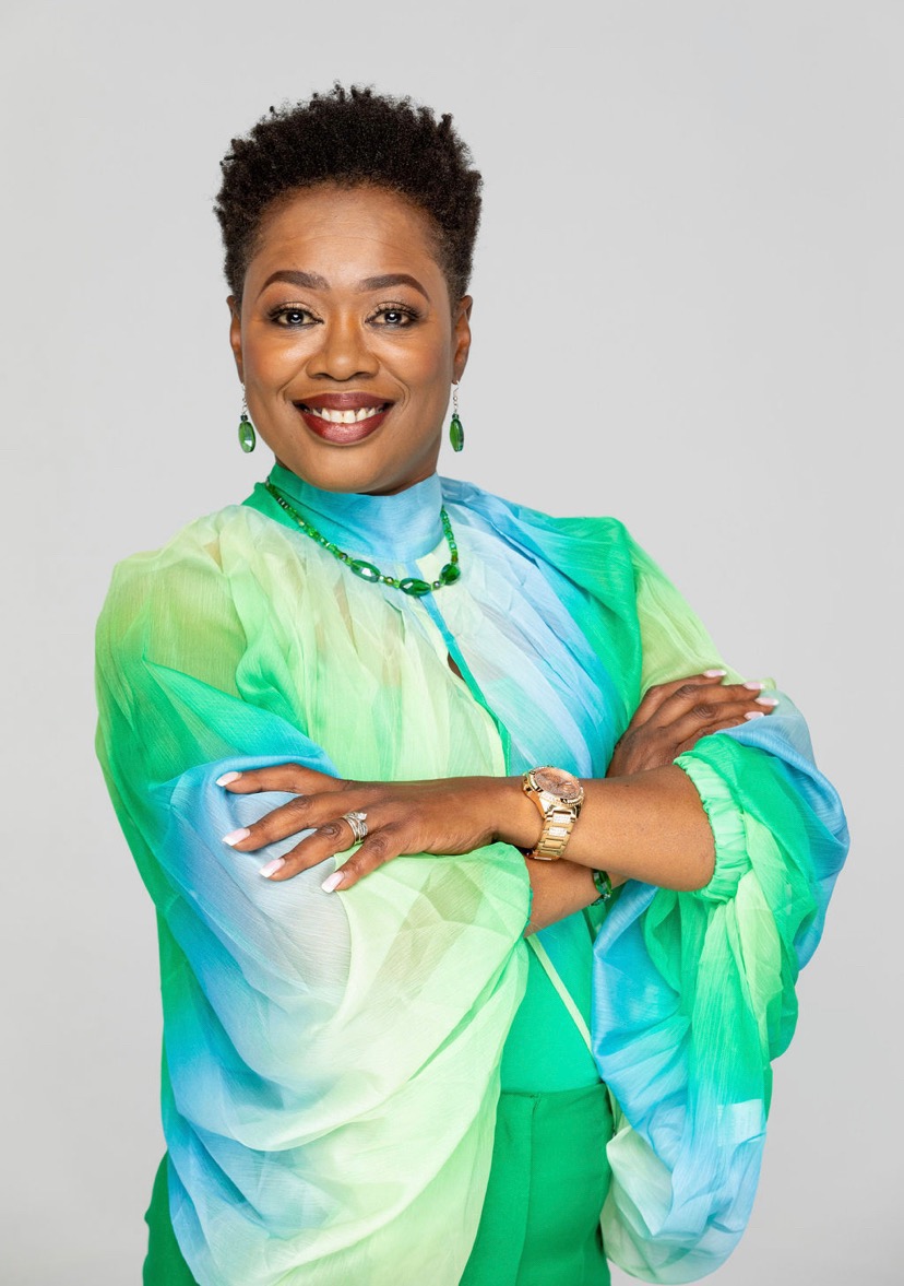 Smiling woman with short curly hair wearing a colorful green and blue dress, jewelry, and a watch, crossing arms.