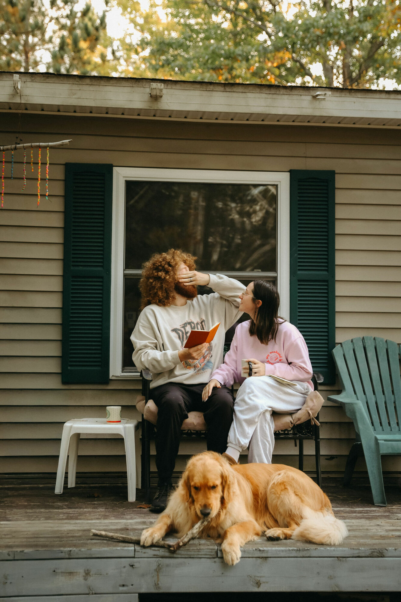 Two women sit on a porch in front of a window with green shutters, with a golden retriever lying on the deck.