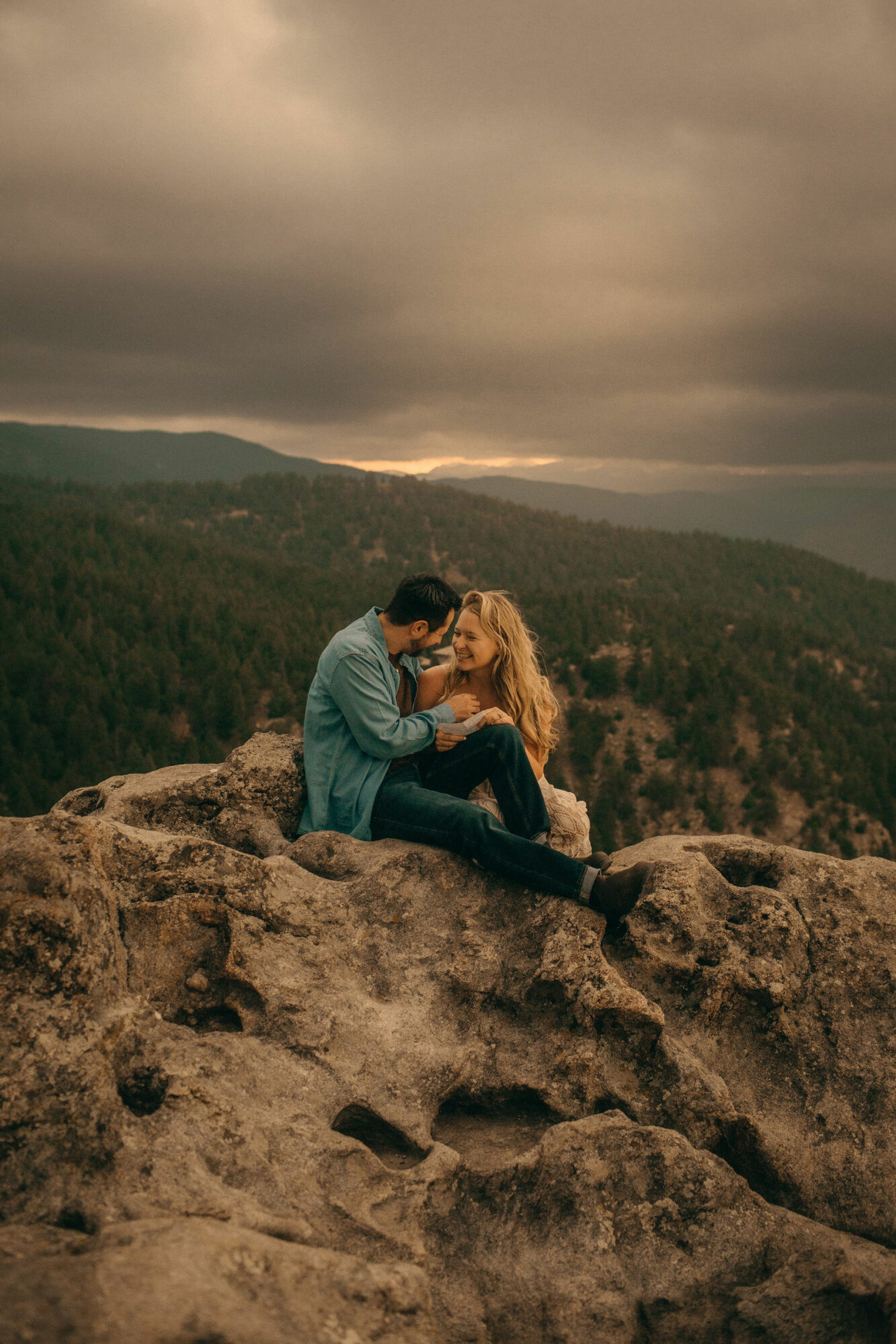 Two people sitting on a large rock, embracing with a landscape of trees and mountains in the background under cloudy sky.