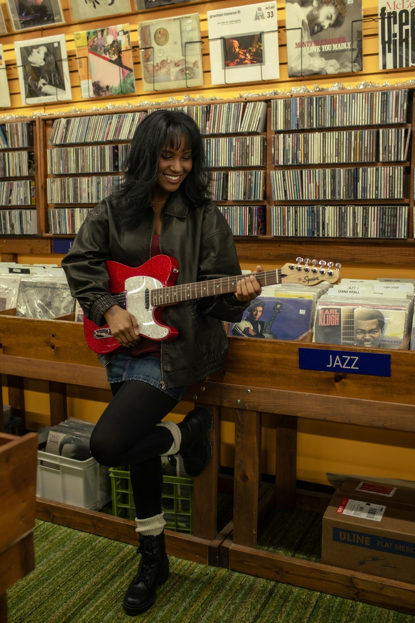 Woman with dark hair playing red and white electric guitar in record store, shelves of vinyl records behind her.