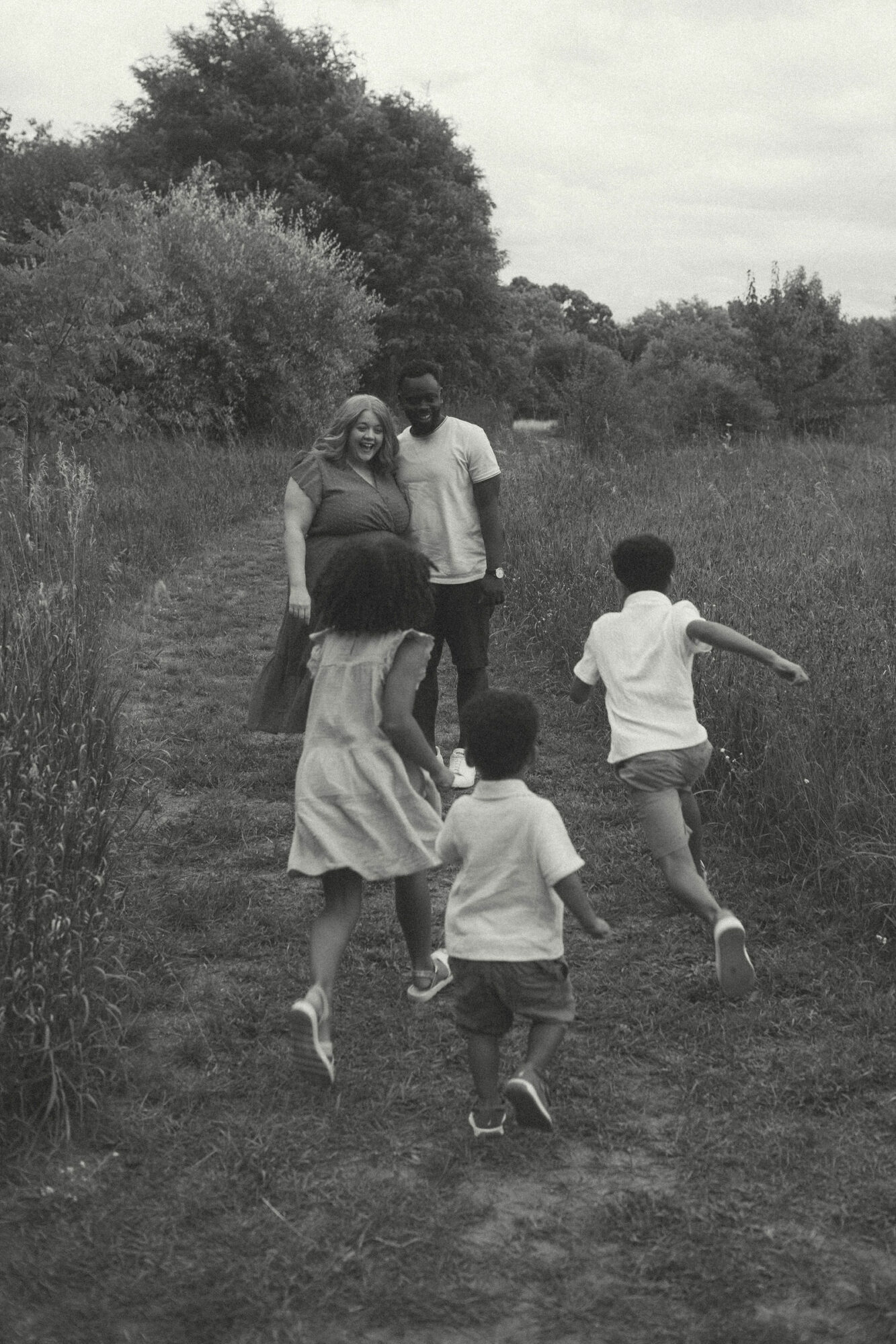 Group of five people, including children and adults, walking outdoors on grass with trees in background.