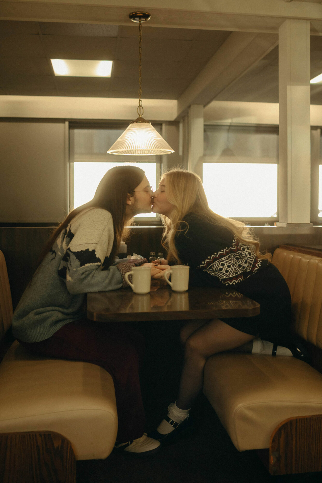 Two women sit at a table in a cafe, leaning towards each other with cups in front of them.