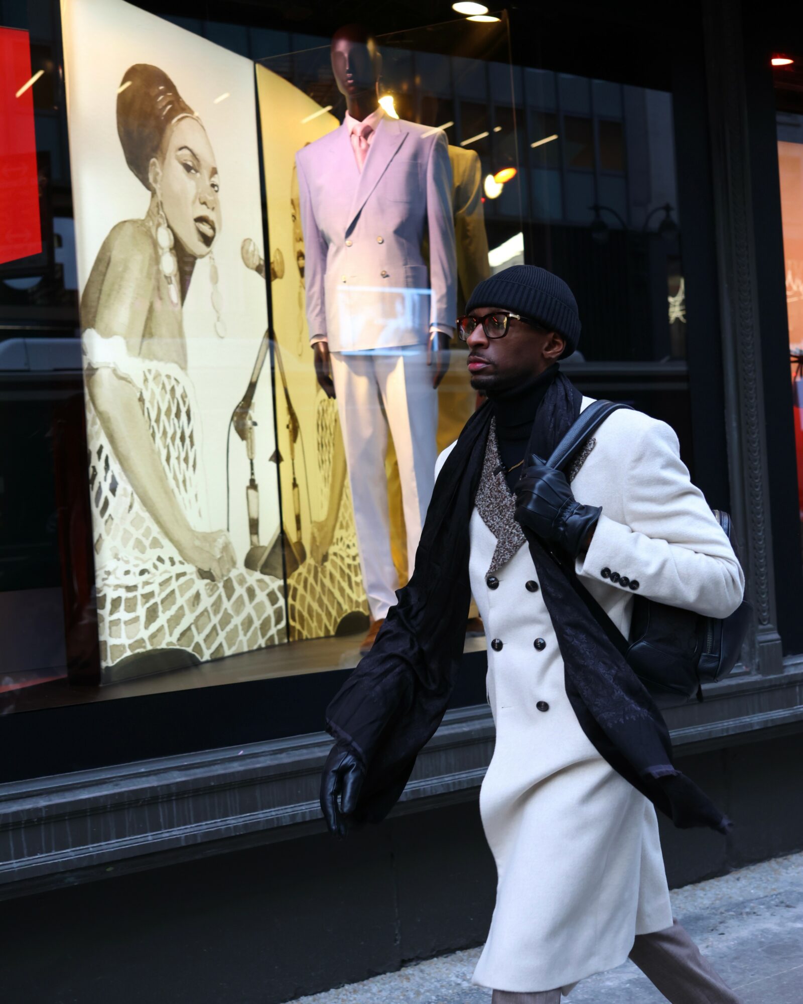 Man in white coat and black gloves walking past a store window with fashion display and large portrait of woman.