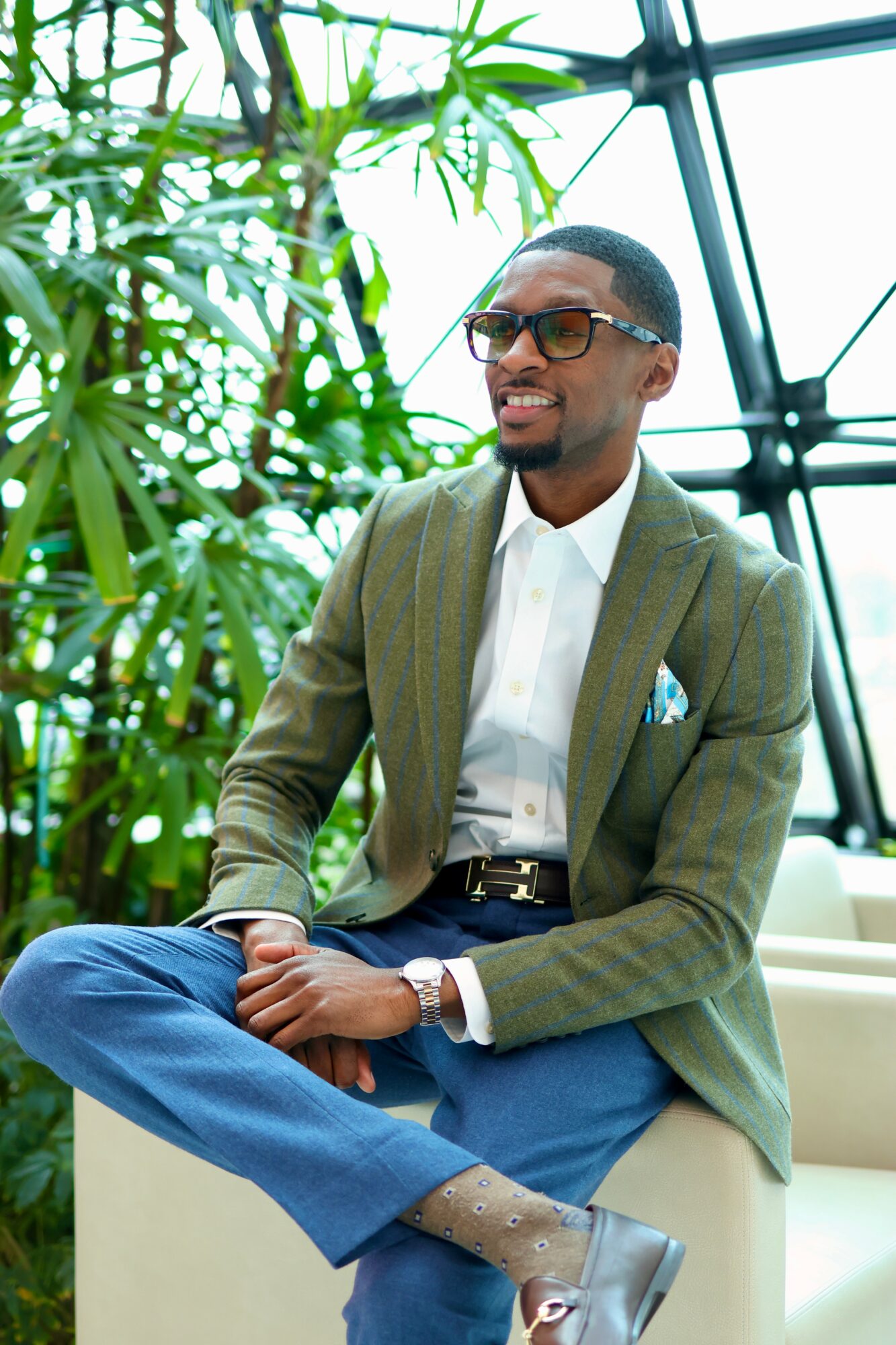 Smiling man in glasses and suit jacket sitting with crossed legs in a greenhouse setting.
