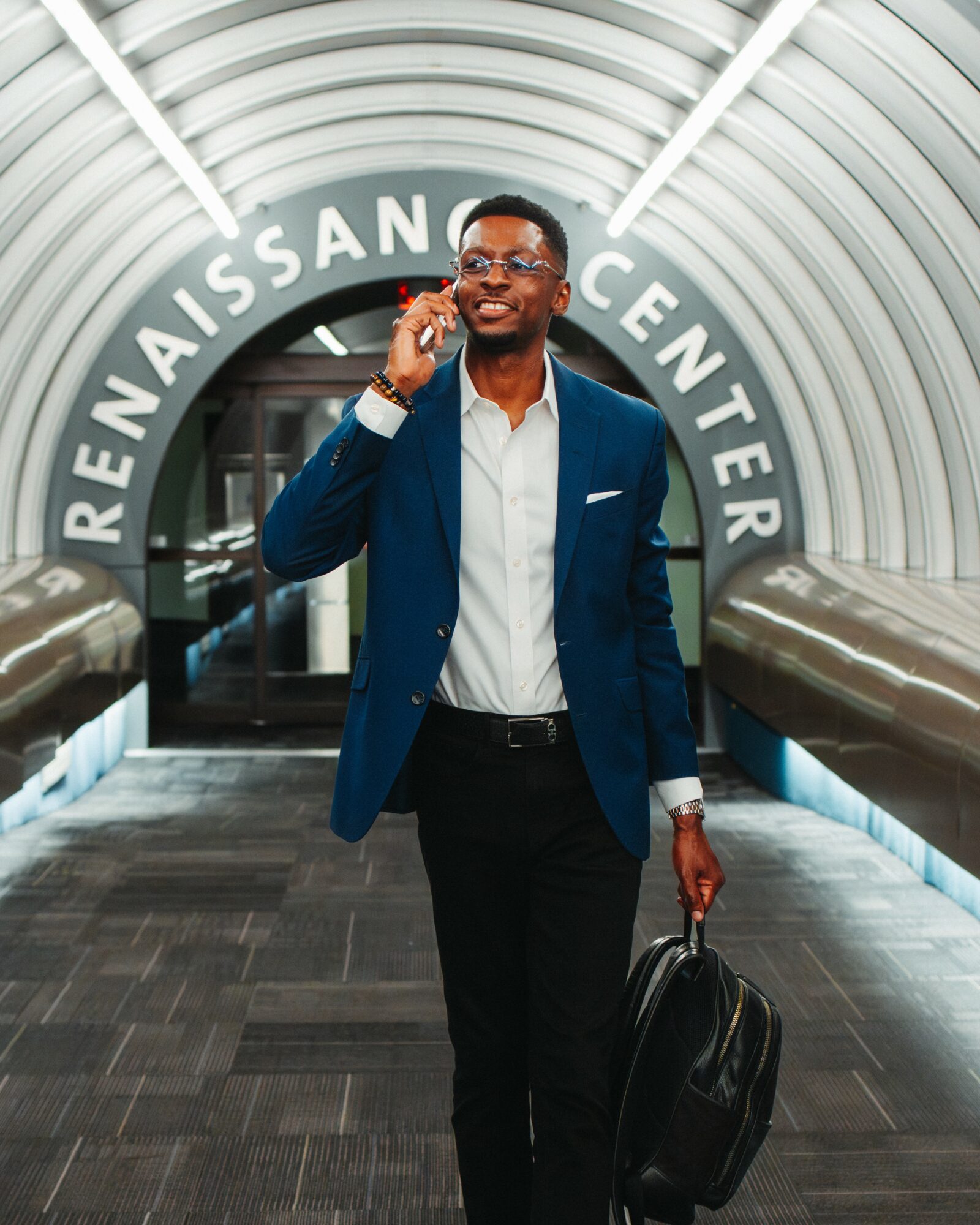 Man in blue blazer and glasses walking through a modern tunnel with curved ceiling, carrying a backpack and talking on phone.