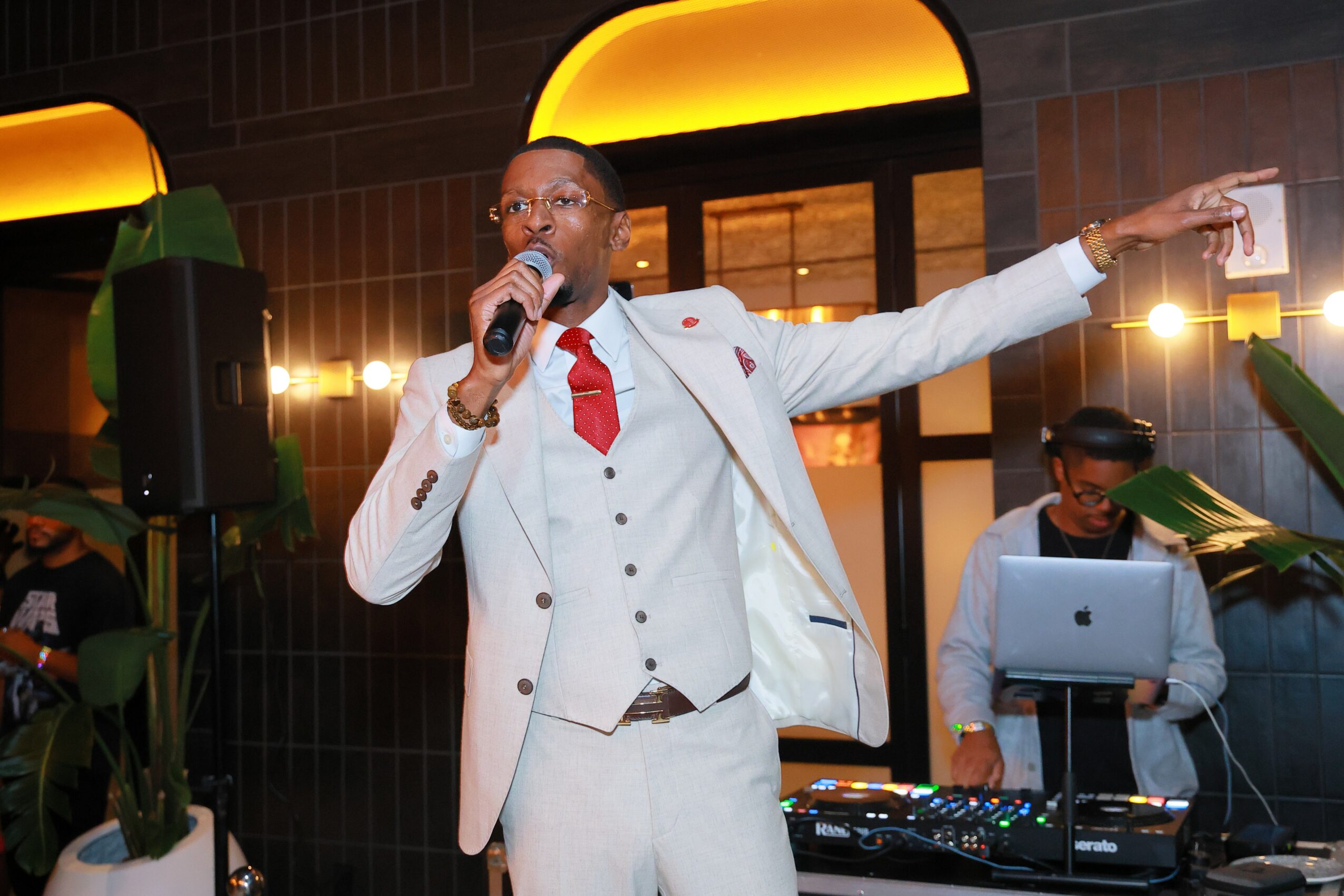 Man in white suit with red tie speaking into microphone, gesturing with arm extended, in indoor setting with people and equipment.